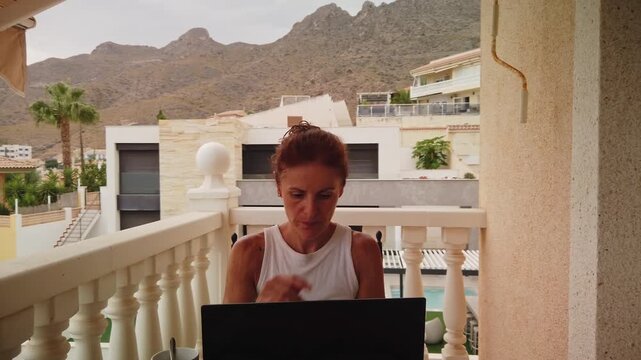Medium shot of a middle-aged woman working focused on her laptop outdoors on a balcony terrace. Scenic coast mountains background, coffee mug, and remote lifestyle vibe.