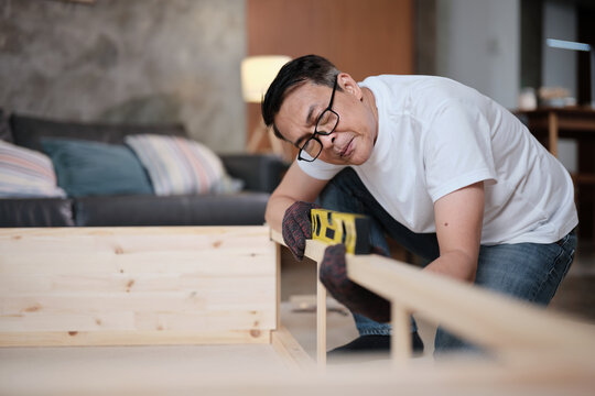 Asian mature man determinedly assembling wooden furniture with DIY tools, checking work precision with spirit&nbsp;level, home improvement, craft installation, hobby carpentry workshop, active lifestyle.