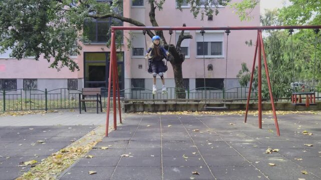 A wide static shot captures a preteen girl wearing a bright blue safety helmet, knee pads, and roller skates as she swings forward on a metal swing set at a residential playground in Hungary.