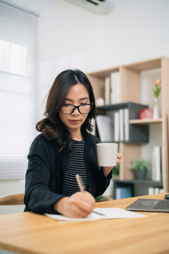 Focused young woman wearing glasses holding coffee cup and writing notes at wooden desk in modern home office interior
