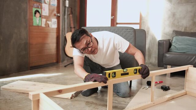 Asian mature man self-assembling wooden furniture with DIY tools, checking work precision with spirit&nbsp;level, home improvement, craft installation, hobby carpentry workshop, handyman active lifestyle.