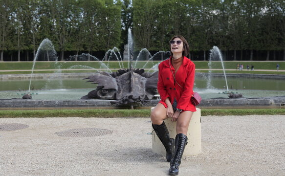 An Asian woman wearing a red jacket, brown turtleneck, and knee-high boots sits on a stone post smiling near a large ornamental pond