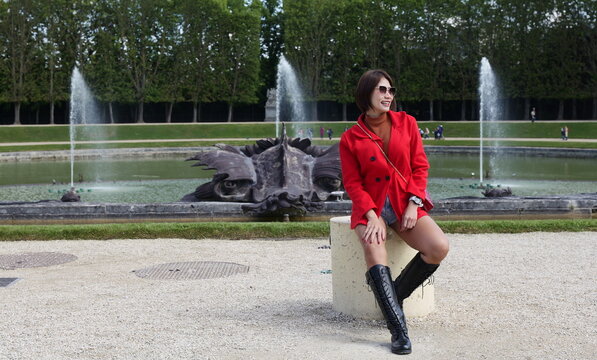 An Asian woman wearing a red jacket, brown turtleneck, and knee-high boots sits on a stone post smiling near a large park fountain basin