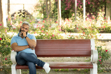 Fototapeta premium Depression, thinking, Upset lonely homeless elderly old Caucasian indian man in garden sitting on bench with depression holds head in hands. Sad, worried senior male with thoughtful face expression