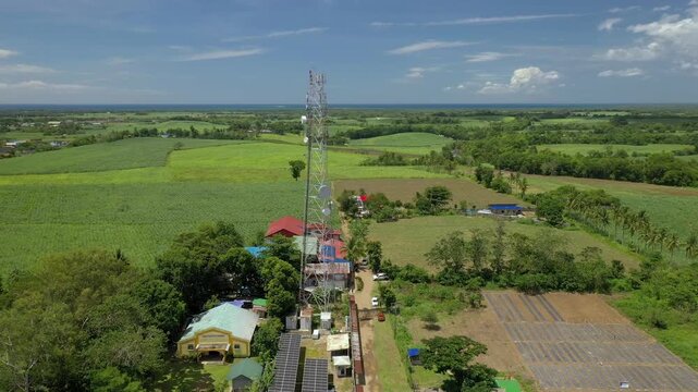 Aerial drone shot panning left and orbiting at 40 meters altitude showing a cell site communication tower with an equipment cabinet. Agricultural land stretches across the background behind the teleco