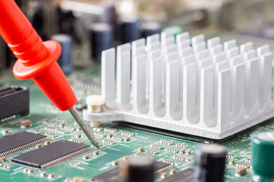 Red multimeter probe testing electronic components on a green circuit board with a silver heatsink