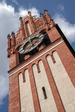 Historic red brick clock tower viewed from below against a bright blue sky with soft white clouds, dramatic low angle architectural perspective, ornate facade with decorative brickwork, arched windows