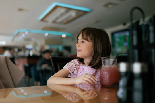 Cute Little Girl Sitting Inside Restaurant Near Healthy Fruit Smoothie. Cheerful young child sits at wooden table watching indoor cafe interior 