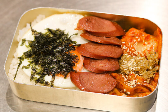 Closeup view of traditional Korean retro lunchbox (dosirak) served in a tin container with rice, fried egg, seaweed flakes, sliced sausage, fish cakes, and kimchi.