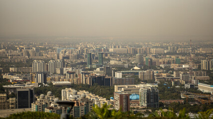Fototapeta premium Wide panoramic view of Almaty, Kazakhstan, showing a dense urban landscape with modern skyscrapers, residential buildings, business centers, parks and city infrastructure under a hazy sky.