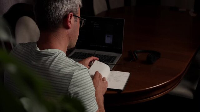 Freelancer works on laptop while taking notes at home office during evening hours