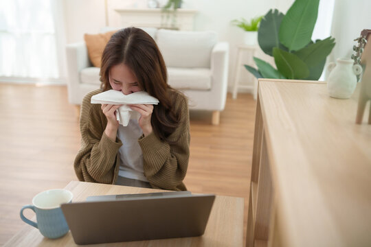Young Asian woman with allergy symptoms uses a tissue while working on a laptop at home, illustrating seasonal illness and sneezing