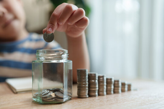Asian Child puts a coin into a glass savings jar beside stacked coins at home, representing financial education, money saving habits and growth in the future goals.