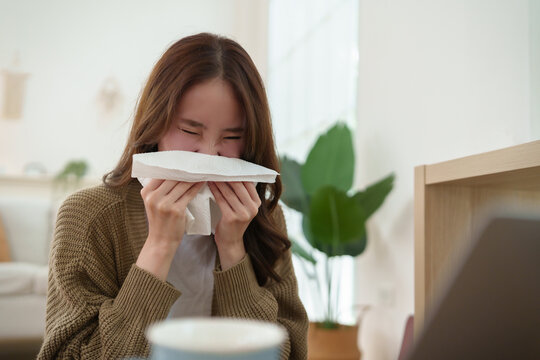 Young Asian woman sneezes into a tissue while studying with a laptop at home, showing allergy symptoms, seasonal illness, health care