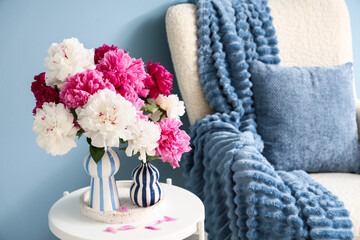 Obraz premium Home interior decor and comfort, fresh peony flowers in striped vases on a side table with a cozy armchair and blue throw blanket in a living room