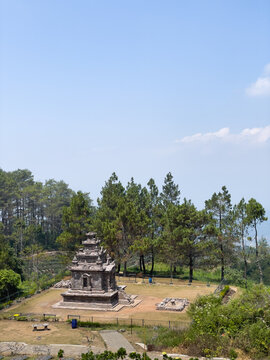 Ancient stone Hindu temple structure sits at Gedong Songo on Mount Ungaran. High-angle shot highlights classic volcanic masonry, pine trees, and clear sky. Ideal for tourism and cultural guides.