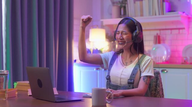 Cheerful young woman wearing a gaming headset, laughing while playing a mobile game on her smartphone, colorful neon game controller sign glowing softly behind her at night.	