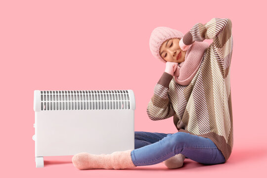 Young Asian woman in winter clothes with radiator sitting on pink background