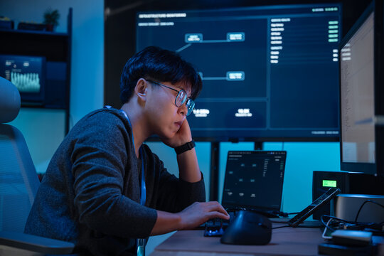 Asian Cybersecurity Analyst Monitoring Network Systems on Multiple Computer Screens During a Late-Night Shift at an Office Workstation
