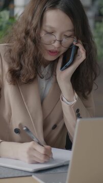 Asian office worker speaking on mobile phone writing information in notebook and using laptop computer. Business and communication concept.