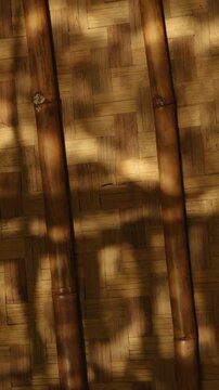 Vertical shot of sunlight and leaf shadows dancing on a woven bamboo wall with bamboo poles, warm rustic aesthetic background.