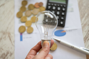 Hand holding an incandescent light bulb over financial documents with coins, a calculator, and a pen, symbolizing rising energy prices and utility expenses © Nanci