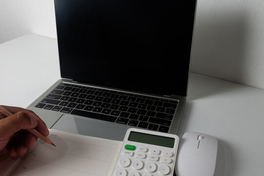 Close up of a hand holding a pencil over a planner with a laptop, calculator, and mouse on a desk. Concept of financial planning, accounting, budget schedule, and work from home with a blank screen.