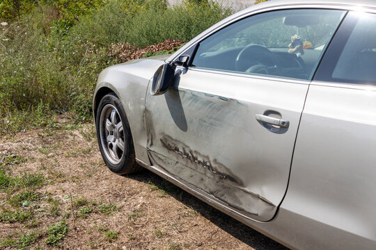 Silver car door and broken side mirror heavily damaged after a traffic accident collision, showing deep scratches, dents, and scuffs in bright outdoor daylight. Car crash
