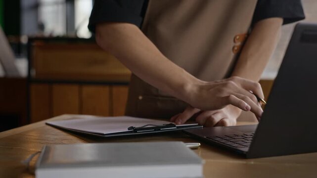 Barista typing on laptop and writing notes on clipboard in coffee shop. Footage works well for small business accounting, cafe management, and inventory commercials