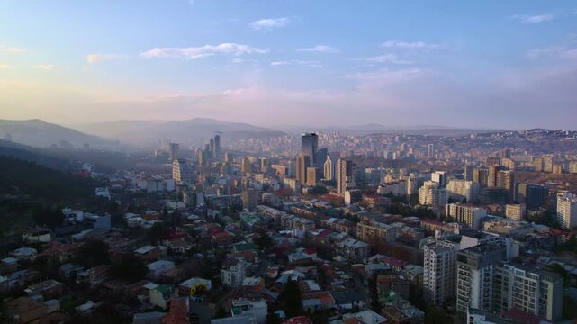 Mountain city panorama moves closer during a gradual drone approach over red and gray rooftops with mid rise blocks modern towers a dark wooded slope and widespread housing beyond