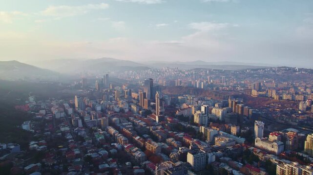 Tbilisi high rises recede through a slow aerial pullback above low roofed neighborhoods linear apartment rows and winter trees with dense housing climbing hills below a pale sky