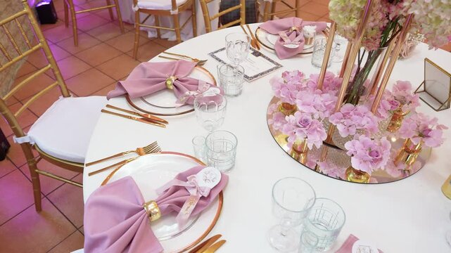 Stylish wedding reception table set with gold cutlery, blush napkins and glassware, featuring a mirrored centerpiece with pink flowers under soft, ambient lighting for a romantic mood.