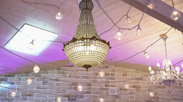 Close-up shot of a grand crystal chandelier in a warm brick room, festooned with glowing string lights. Soft purple uplighting enhances the inviting, festive ambiance for events.