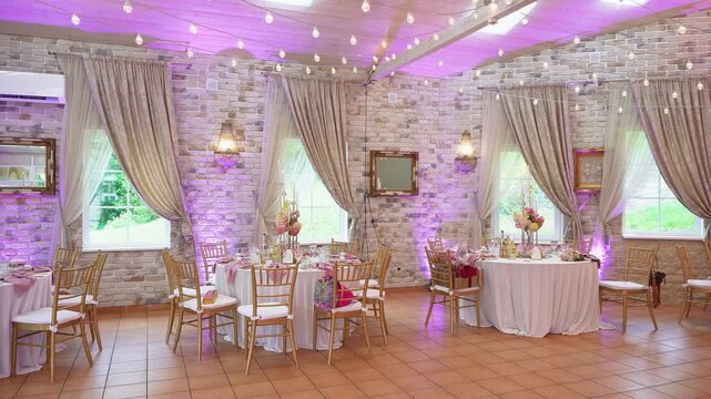 Beautifully decorated wedding reception hall with draped curtains, string lights, floral centerpieces, and gold chairs. Soft purple uplighting creates a magical, festive atmosphere in an empty event