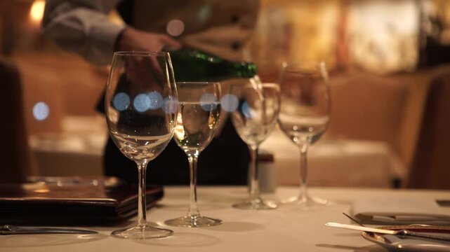 Waiter pouring still water into crystal glasses on white tablecloth in elegant upscale restaurant setting.