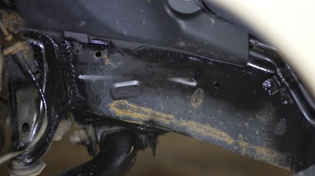 Man spraying black protective anti-corrosion coating onto car chassis part.