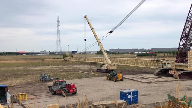 Large crane and heavy construction equipment at site preparation for assembly of massive lattice jib on open field.