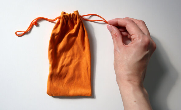 A person is using their hand to hold an orange drawstring bag. The bag is made of soft material and is being secured at the top. The background is plain and white.