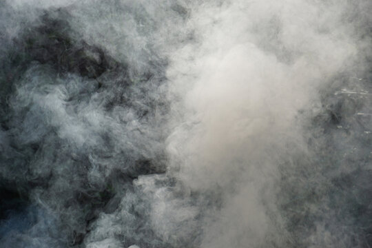 A close-up image of thick white smoke rising, billowing, and swirling unpredictably against a dark, shadowy background, creating a dramatic and abstract texture.