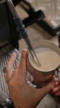 Vertical video of barista steaming milk in stainless steel pitcher with espresso machine steam wand. Ideal for cafe branding, coffee marketing, barista training