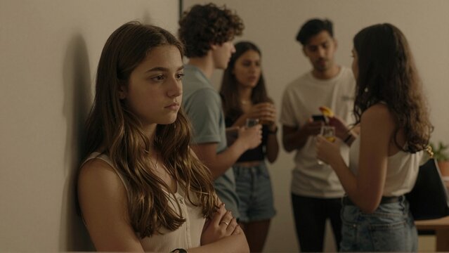 Withdrawn teenage girl stands apart as friends chat with drinks at indoor party