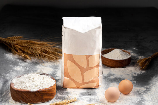 White paper flour package mockup with wheat ears eggs and flour in wooden bowls on dark concrete background