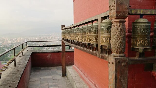 Row of prayer wheels spinning on a balcony overlooking a city, traditional spiritual objects.