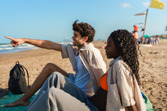 Happy young diverse couple relaxing on sandy beach pointing, sharing a moment