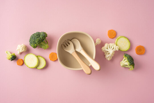 Baby's first feeding setup with silicone bowl, wooden utensils and fresh vegetables for healthy eating on light pastel pink table