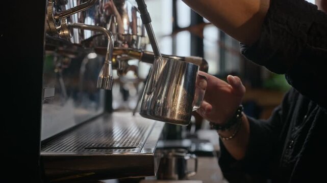 Caucasian young adult barista steaming milk with espresso machine steam wand into stainless pitcher and cleaning it with cloth. Clip suits coffee shop commercials and barista training courses
