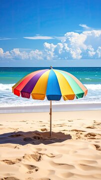 A vibrant beach umbrella stands on the sand under a bright blue sky with fluffy clouds and turquoise ocean waves.