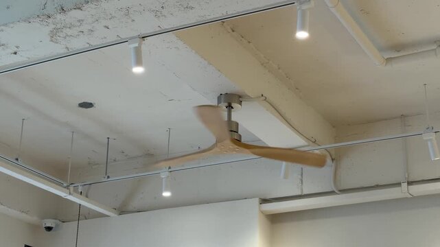 Modern wooden ceiling fan spinning on a white industrial exposed concrete ceiling with track lighting