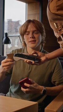 Vertical shot of Caucasian young man paying with credit card on POS terminal and drinking coffee in cafe. Perfect for mobile banking, digital fintech campaigns and coffee shop promo