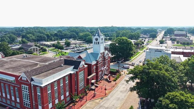Red brick courthouse with white clock tower anchors small town square in Atlanta, Georgia, cars drive tree lined main street past historic storefronts under bright sun. Aerial drone wide shot